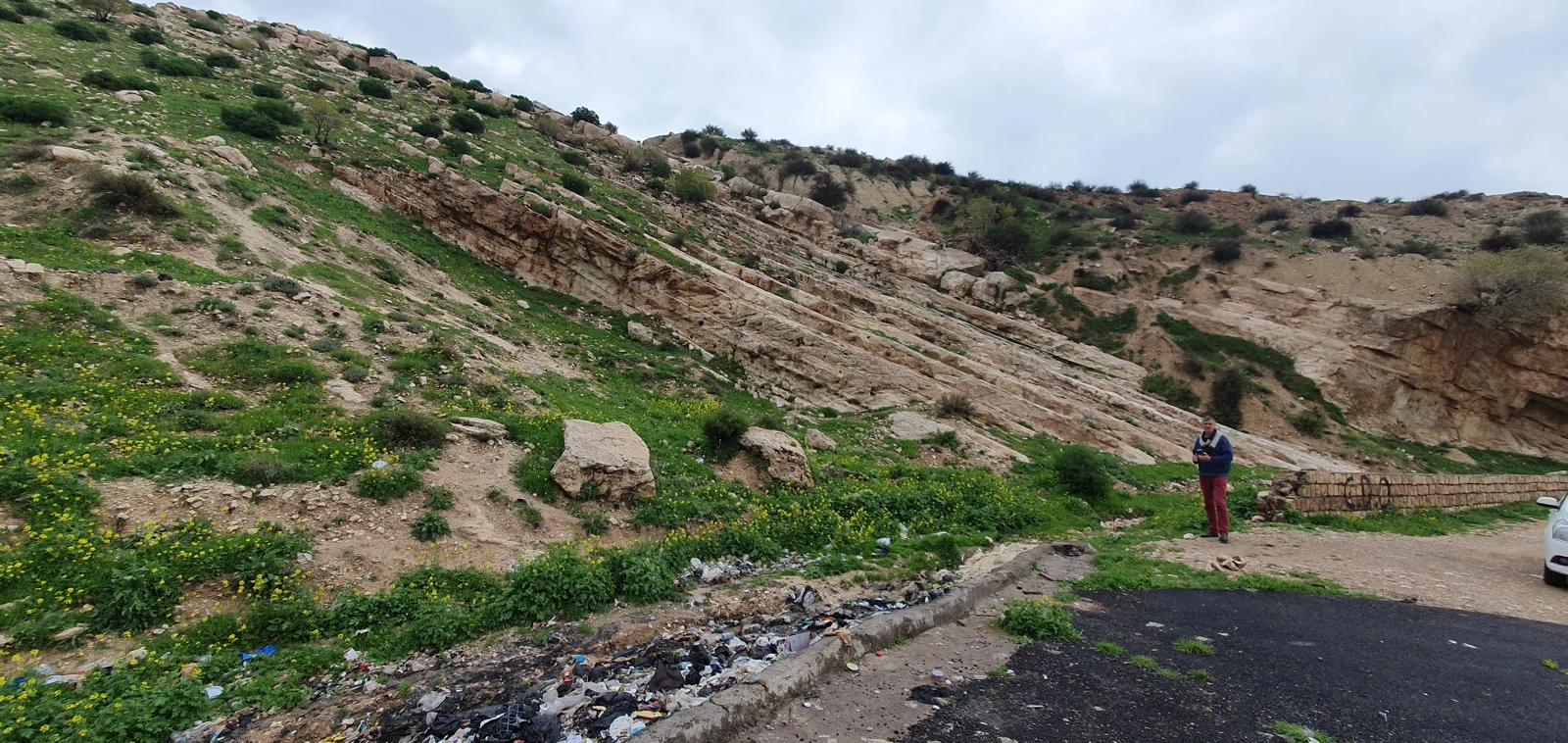 Rubbish dumped beside a road below green, rocky hills.